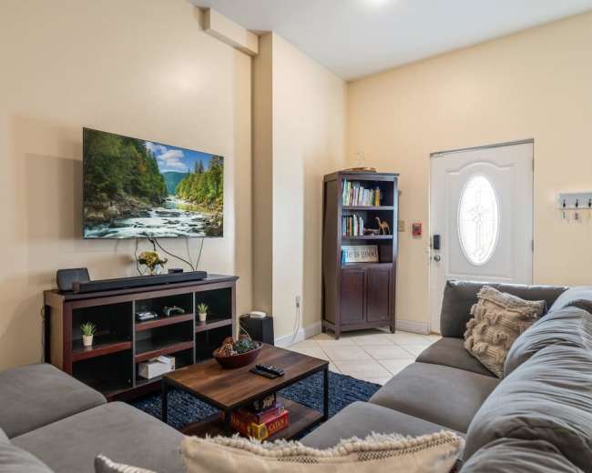 A living room features a large TV mounted on the wall opposite a gray sectional sofa, with a wooden coffee table and a door leading outside.