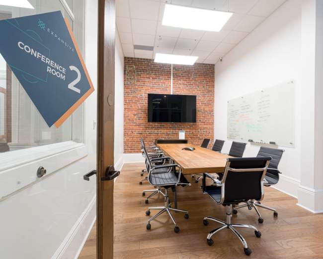 A conference room featuring a large wooden table, several black chairs, a television screen on a brick wall, and a whiteboard for presentations.