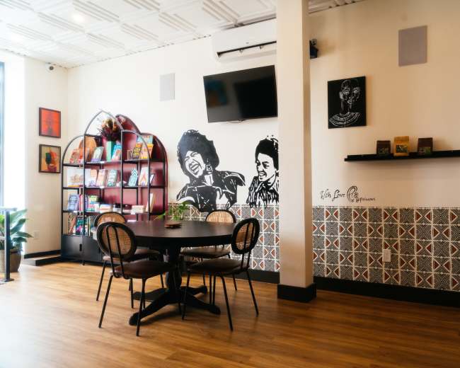 The image shows a cozy interior of a café featuring a round table, bookshelves against the wall, and framed artwork of people, alongside a patterned tile design.