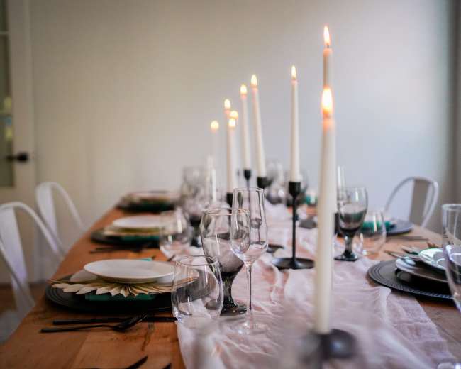 A long wooden dining table is set with plates, glasses, and lit candles arranged in a row.