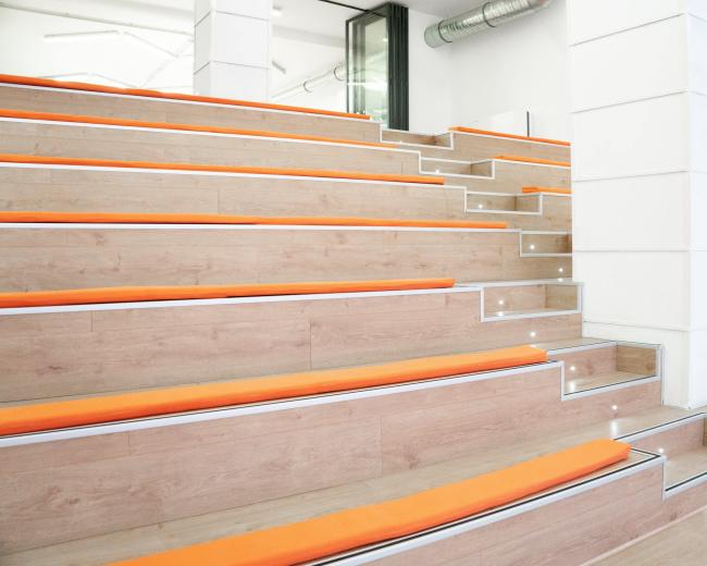 The image shows a modern auditorium with stepped seating lined with orange cushions.