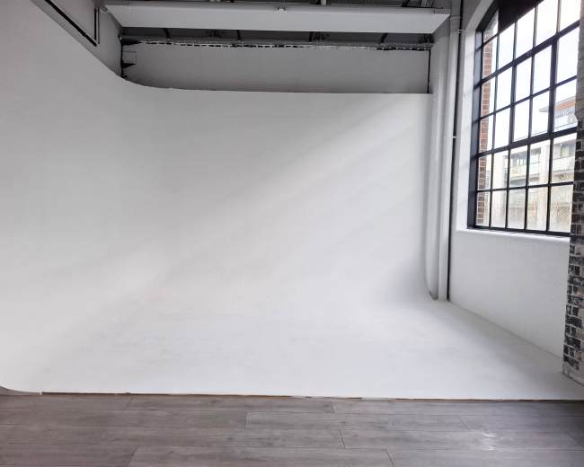 The image shows a spacious, empty photo studio with a white seamless backdrop and large windows allowing natural light to enter.