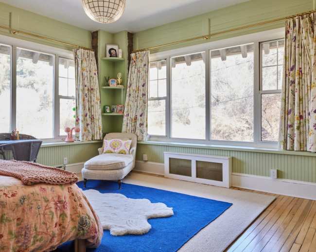 The image shows a softly lit bedroom with large windows, floral curtains, a white rug, and a beige chair next to a small table.