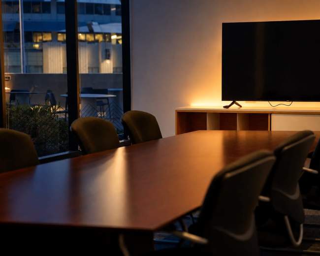 A conference room features a long wooden table surrounded by black chairs, with a television mounted on a wall illuminated by soft lighting.