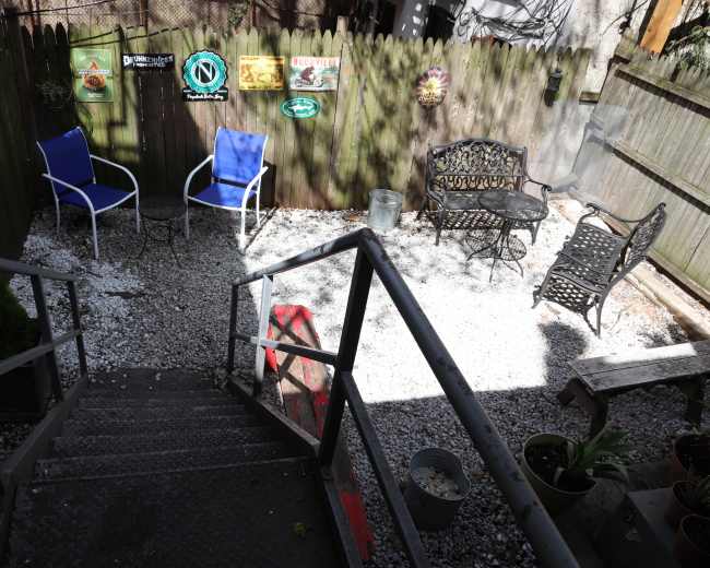 The image shows a gravel patio area with two blue chairs and a wrought iron table set against a wooden fence adorned with various signs.