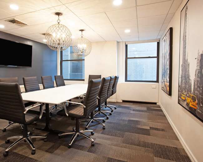 A modern conference room features a long white table surrounded by black swivel chairs, with pendant lighting and a wall-mounted TV.