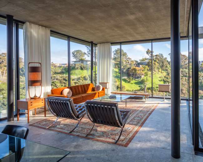 The image shows a modern living space with large glass windows, featuring an orange sofa, two black chairs, and a view of green hills outside.