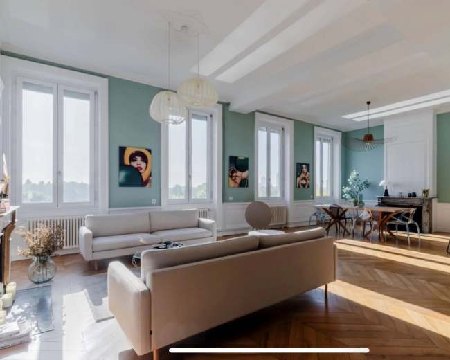 The image shows a modern living room with light green walls, large windows, and stylish furniture, including a sofa and a round chair.