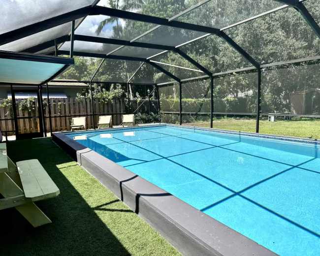 The image shows a rectangular swimming pool enclosed by a mesh screen structure, surrounded by a grassy area and a wooden fence.