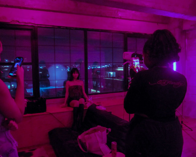 Three people are in a dimly lit room with purple lighting, with one person posing on a bed while the others take photos using their phones.