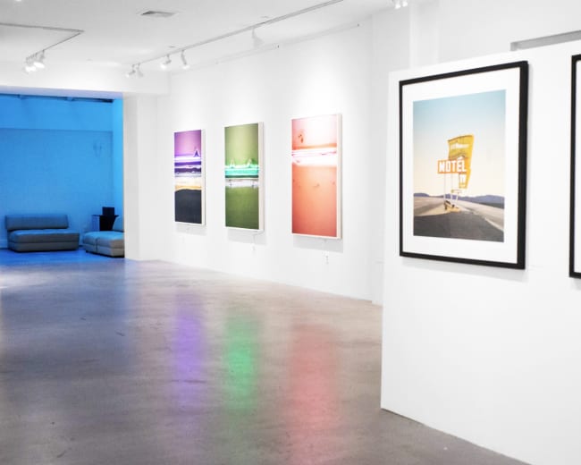 The image shows an art gallery with framed photographs displayed on white walls, leading to a blue-lit area in the background.