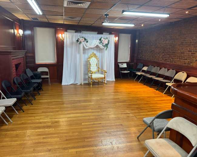The room features a decorative throne in front of a white draped backdrop, surrounded by rows of empty chairs on polished wooden flooring.