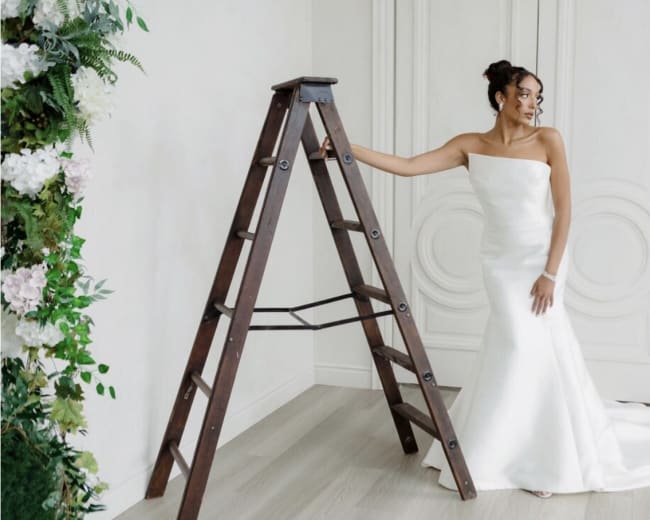 A woman in a strapless white wedding dress stands beside a wooden ladder in a room with light-colored walls and a floral backdrop.