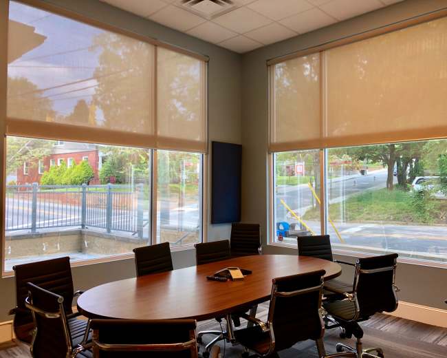 A large conference room features a round wooden table surrounded by black chairs, with two large windows covered by light-colored shades.