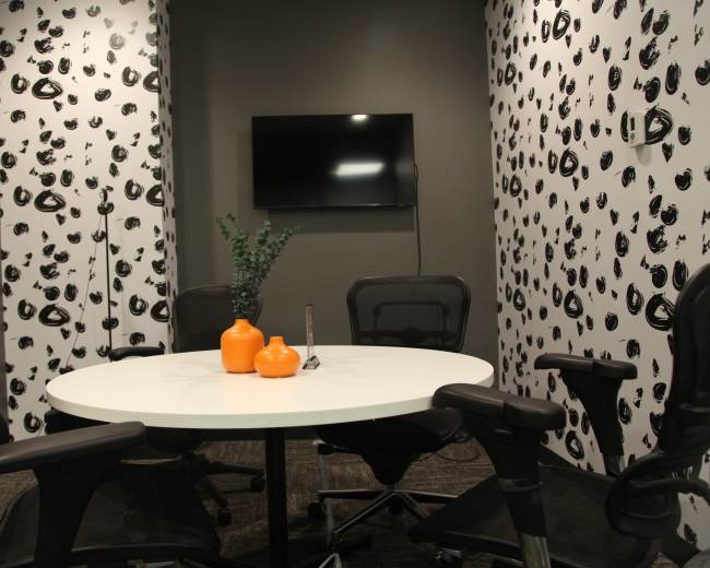 A conference room features a round white table surrounded by black office chairs, with patterned walls and a television mounted on one side.