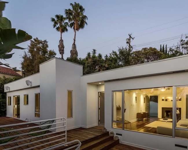 The image depicts a modern single-story house with large windows and a wooden deck, surrounded by palm trees and greenery.