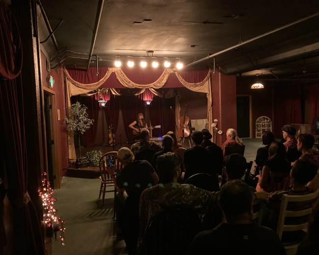 A musician performs with a guitar on stage in a dimly lit venue while an audience sits at tables.
