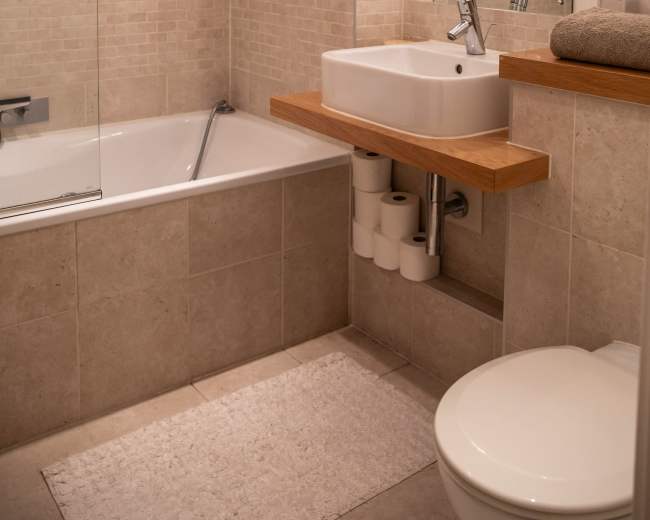 A modern bathroom with a bathtub, a wall-mounted sink on a wooden shelf, a mirror, and neutral-toned tile walls.