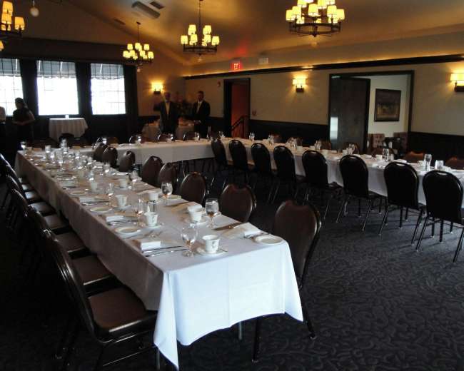 A dining room is set up with several long tables covered in white tablecloths, arranged with dishes, glasses, and utensils.