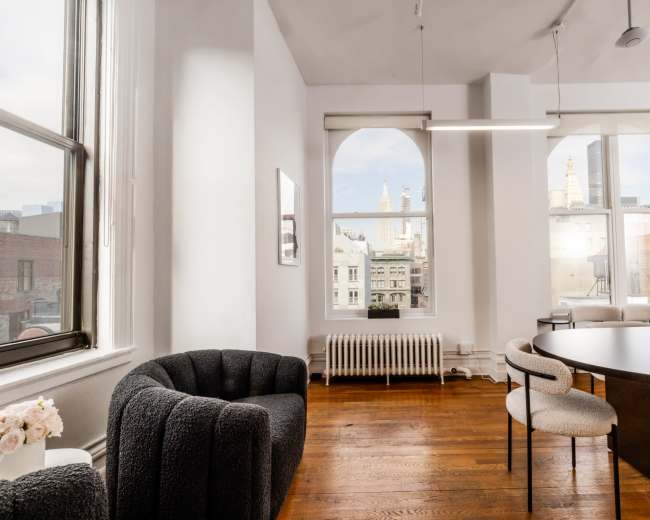 The image shows a modern interior office space with large arched windows overlooking a cityscape, featuring a round table and a cozy armchair.