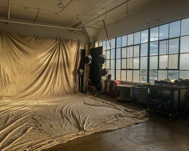 The image shows a photography studio with a wrinkled fabric backdrop, studio lights, and equipment near a large window letting in natural light.