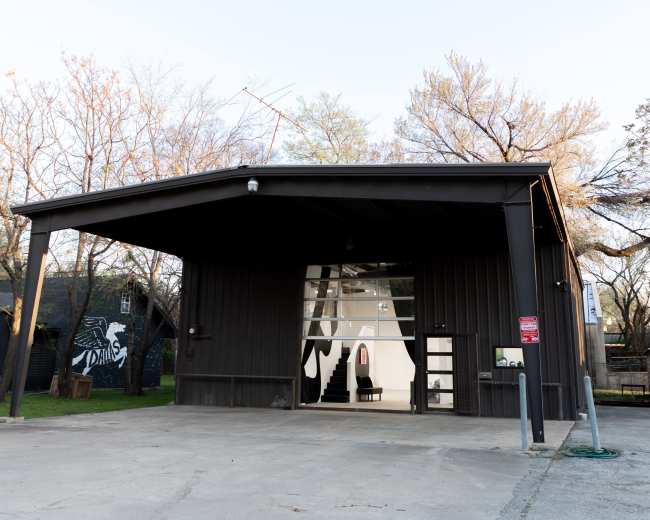 A black metal building with a large front porch and large windows sits on a paved lot, surrounded by bare trees and grassy areas.