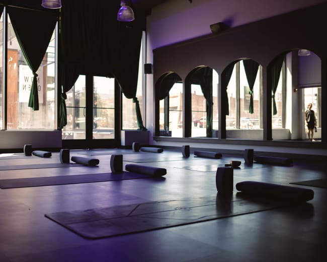 A dimly lit yoga studio features several rolled mats arranged on the floor, with large windows allowing natural light to stream in.