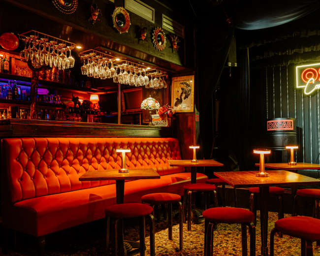 The image shows a bar area with red seating, small tables, and a neon sign that reads "OSCAR," set in a dimly lit, stylish interior.