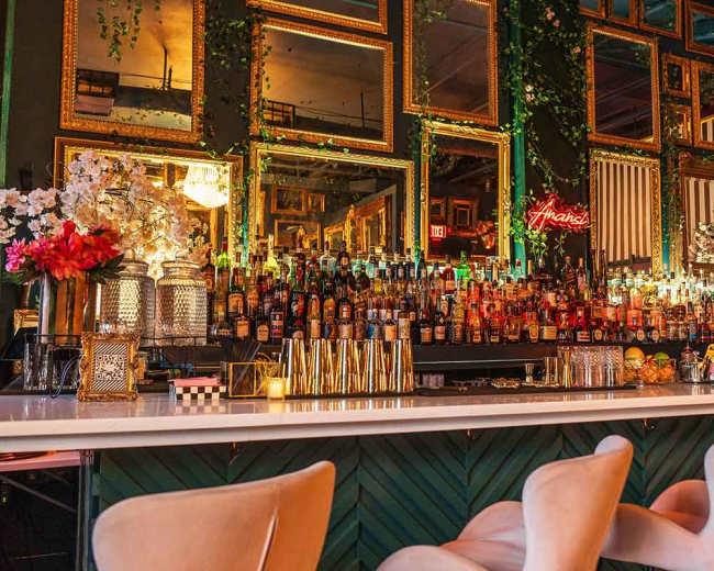 The image shows a stylish bar area featuring a long white countertop, an array of colorful bottles organized on shelves behind it, and multiple ornate golden mirrors reflecting the scene.