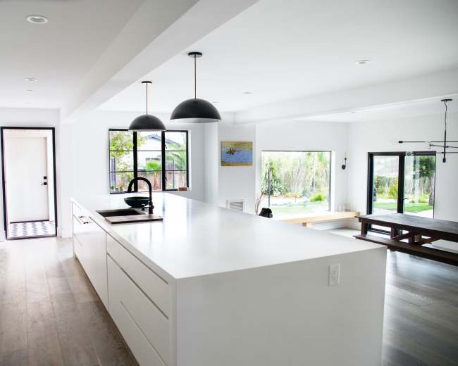 The image shows a modern, minimalist kitchen with a long white countertop and black fixtures, connected to a bright living area featuring large windows.