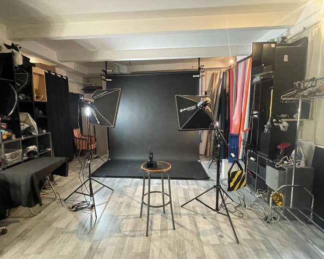 The image shows a photography studio with two softbox lights, a stool in the center, and a black backdrop.
