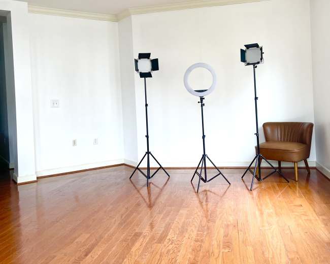 The image shows a spacious, empty room with wooden flooring, featuring three lighting stands and a brown chair against a white wall.