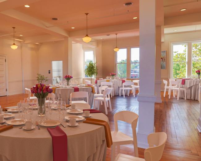A bright event space with several round tables set with white tableware and floral centerpieces, surrounded by white chairs.