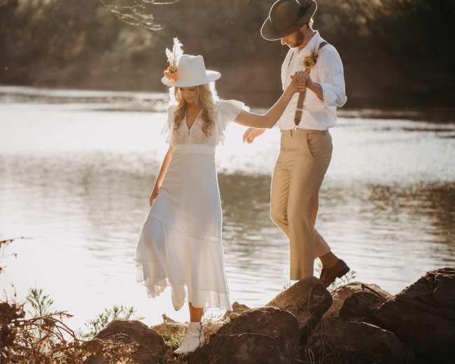 A couple walks hand in hand along the rocky shore of a river, with the sun setting in the background.