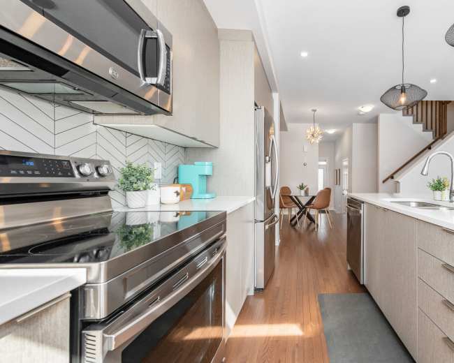 A modern kitchen features stainless steel appliances, a central island with a sink, and light wood cabinetry, leading into a bright dining area.