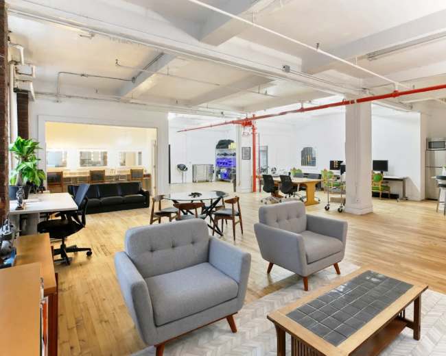 The image shows a modern open office space with wooden floors, several seating areas, and a kitchenette in the background.