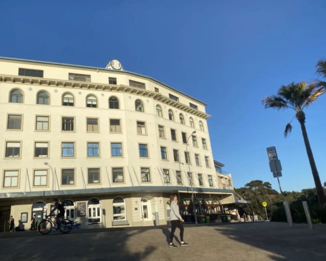 A person walks past a historic building with a clock on the roof, located in a coastal area with palm trees.