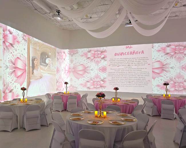 A spacious event room is decorated with floral projections on the walls and set up with round tables covered in white with pink accents.