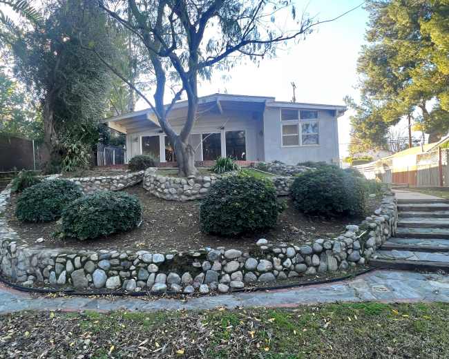 The image shows a house with a stone garden featuring circular shrubbery, surrounded by steps and trees.