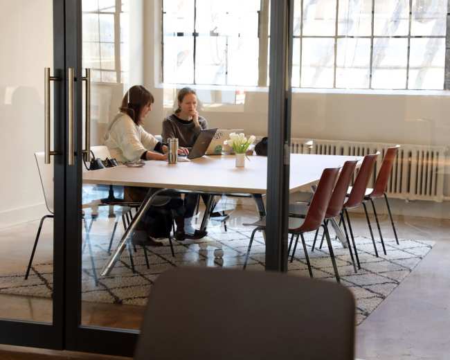 Two women are sitting at a table in a well-lit office, working on laptops with a vase of flowers in the center.