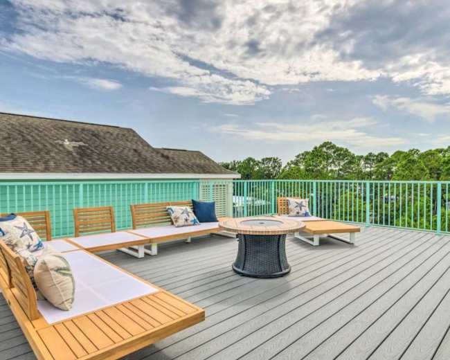 The image shows a spacious outdoor deck with wooden seating around a circular table, surrounded by green trees under a partly cloudy sky.