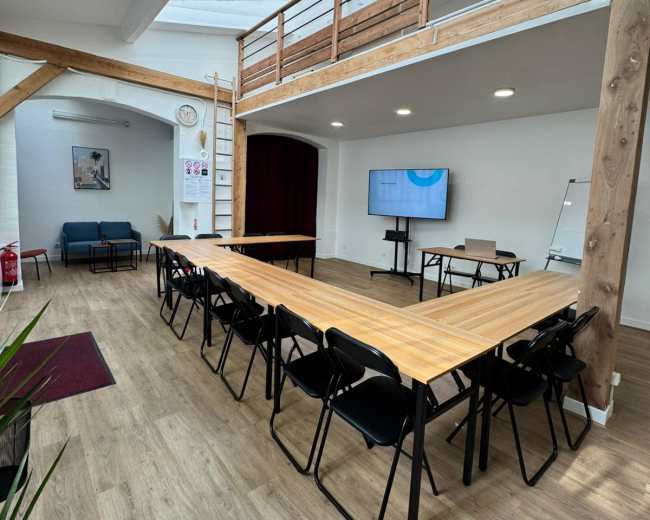 The image shows a spacious, well-lit meeting room with a large wooden table arranged in a V-shape, surrounded by chairs, and featuring a screen on one wall.