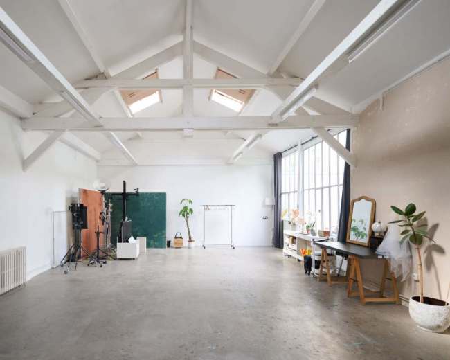 The image shows a spacious, empty room with high ceilings, large windows, and minimal furniture, including a desk and some studio equipment.