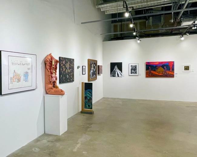 The image shows an art gallery with various pieces of artwork displayed on white walls, including paintings and a sculpture.