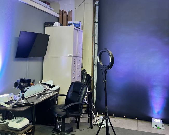 A workspace with a desk and chair, a monitor, and a ring light set up in front of a dark backdrop.