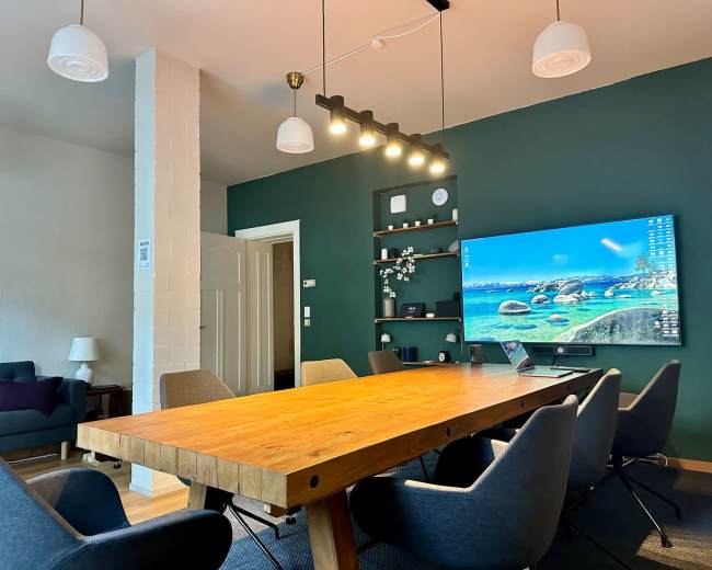 The image shows a modern conference room with a large wooden table surrounded by chairs, a mounted television displaying a scenic background, and a dark green accent wall.