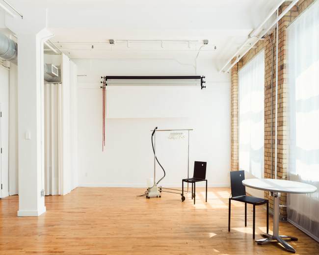 The image shows a minimalist interior space with wooden flooring, large windows, a clothing rack, a chair, and a small table.