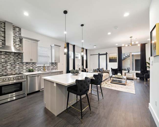 The image shows a modern kitchen and living area featuring stainless steel appliances, a stone backsplash, and a central island with bar seating.