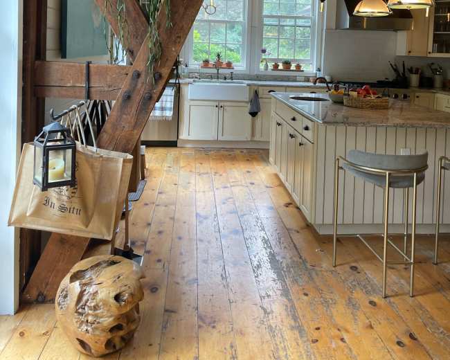 A rustic kitchen features exposed wooden beams, a central island, and large windows with greenery outside.