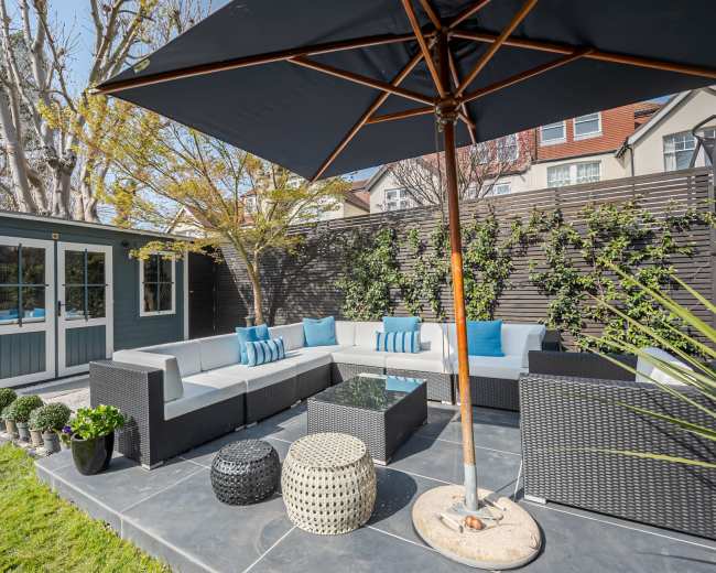 The image depicts a cozy outdoor seating area with a sectional sofa, an umbrella, and a garden shed surrounded by greenery.
